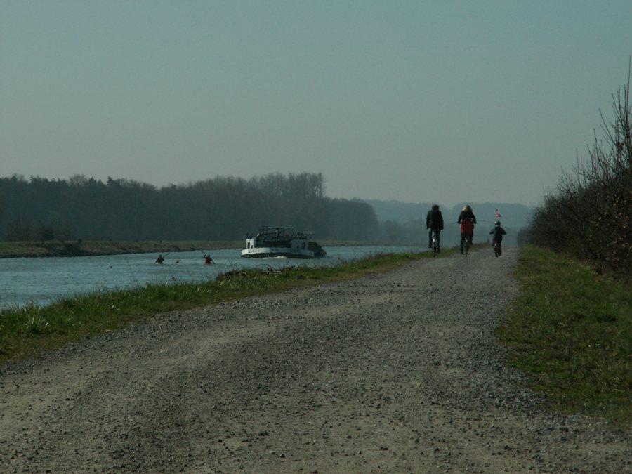 Drei Radfahrer auf einem Schotterweg am Fluss mit Ausflugsschiff und Kanufahrern Drei Radfahrer auf einem Schotterweg am Fluss mit Ausflugsschiff und Kanufahrern